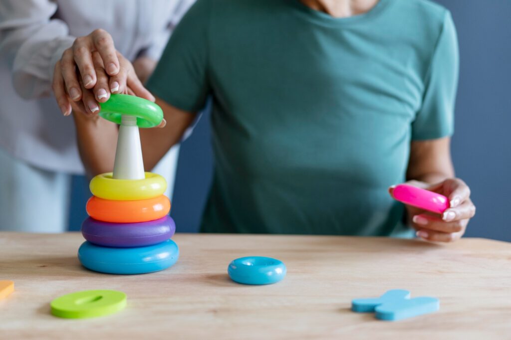 A imagem mostra uma pessoa interagindo com um brinquedo educativo de anéis coloridos. A pessoa está empilhando os anéis em ordem de tamanho, começando pelo maior e indo até o menor. Os anéis são de cores vibrantes: verde, amarelo, laranja, roxo e azul. A pessoa está usando uma mão para empilhar os anéis e a outra mão está segurando um anel azul. O brinquedo está sobre uma superfície de madeira clara, e ao fundo há uma parede azul. A pessoa está vestindo uma camiseta verde e tem unhas pintadas de branco.