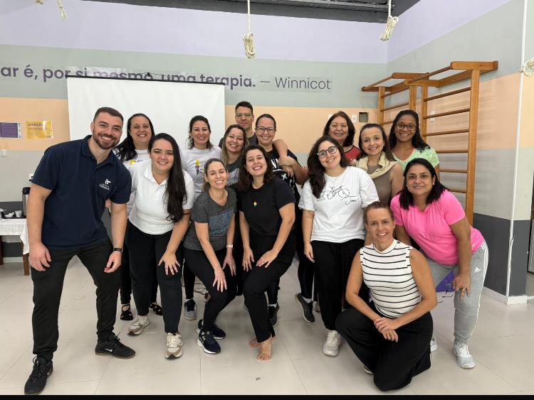 Grande grupo de pessoas sorrindo em uma sala de terapia, com citação de Winnicott no fundo.