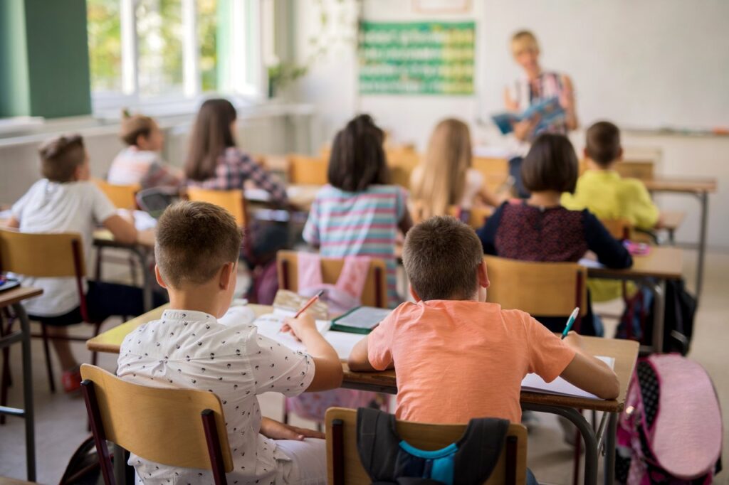 Sala de aula vista de trás: alunos sentados em duplas acompanham a professora, que explica o conteúdo em pé à frente da turma
