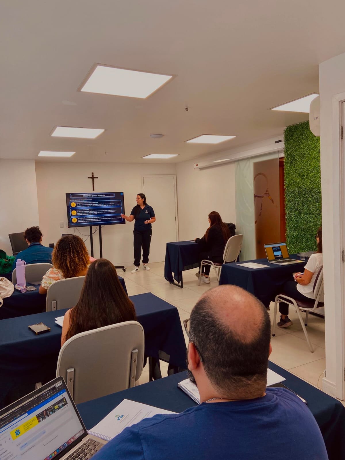 Sala de aula do Instituto Celebra durante a turma de Recertificação: a instrutora aponta para um slide com diretrizes para a prática enquanto profissionais acompanham sentados em mesas com apostilas e notebooks.